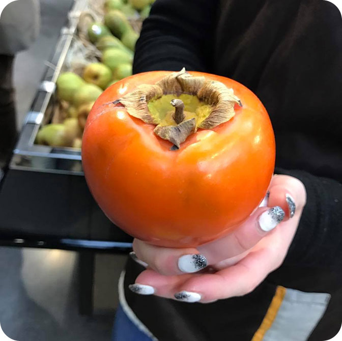 Gros plan sur un kaki tenu par une personne, avec des fruits flous en arrière-plan.